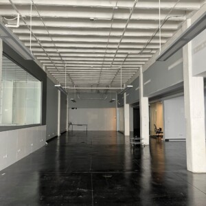Interior view of a commercial space prior to renovation, showing a long corridor with black flooring, white ceiling beams, exposed lighting conduit, and large interior windows on the left. Minimal furnishings are present, indicating the space is in a pre-remodel state.