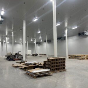 Northstar Pickle - Finished Goods Cooler "Spacious interior of an industrial cold storage or finished goods facility with tall white support columns, high ceilings, and wall-mounted cooling units. Pallets of materials, cardboard, and processing equipment are positioned throughout the clean, concrete-floored space.