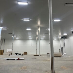 Northstar Pickle Finished Goods Cooler Interior of a large industrial cooler facility with white insulated walls, polished concrete floor, bright ceiling-mounted LED lighting, and several vertical support columns. Construction materials and equipment are scattered around the space, indicating ongoing work.