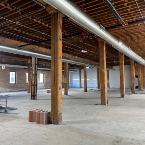 Wide-angle view of a partially completed commercial interior showing exposed wooden beams, overhead metal ductwork, and unfinished concrete floors. Several windows line the brick wall, and construction materials such as a rolling tool cart and cardboard boxes are scattered throughout the open space.
