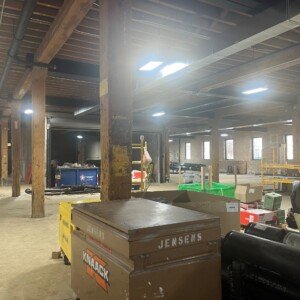 "Interior view of an active construction site in a large, open commercial space with exposed brick walls, wooden support beams, and overhead ductwork. Construction materials, toolboxes labeled 'KNAACK' and 'JENSEN,' scaffolding, and cardboard boxes are scattered throughout the site.