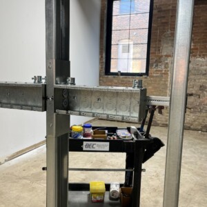 Close-up of in-progress electrical installation inside a construction site. Exposed metal framing and mounted electrical boxes are shown, with a cart labeled 'Benson Electrical Contracting' holding tools and supplies in the background. A brick wall and window are visible behind the setup.