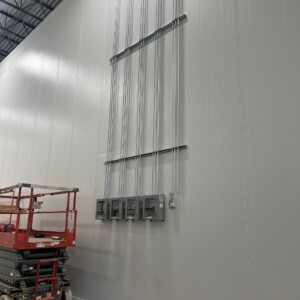 Northstar Pickle Electrical installation inside a commercial facility featuring multiple vertical conduit lines connected to electrical panel boxes mounted high on a tall, light gray wall. A red scissor lift is parked nearby, indicating ongoing work.