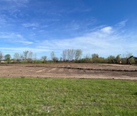 Open grassy construction site under a blue sky with scattered trees in the background.